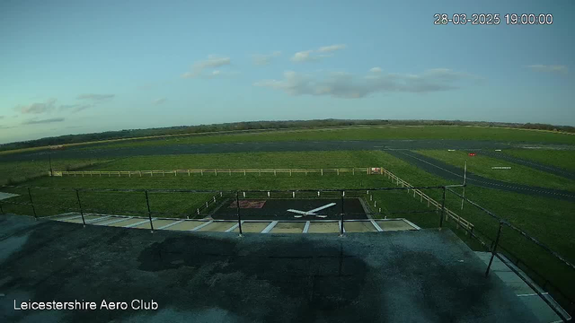 A wide view of an airfield at Leicestershire Aero Club during the evening. The sky is mostly clear with a few clouds. In the foreground, a helicopter landing pad can be seen with a white cross marking. Beyond that, the grassy airfield stretches out, bordered by a wooden fence. On the right, a paved runway is visible, with a red flag marking its end. The scene is serene with no aircraft present.