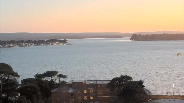A tranquil waterfront scene during sunset. The sky transitions from soft orange to pale blue, while calm water reflects the colors. In the foreground, a cluster of dark green trees lines the shore. A few buildings with lighted windows are visible on the left, along with small boats along the coastline. The distant landscape features hills and more trees, with a gentle horizon where the sky meets the land.