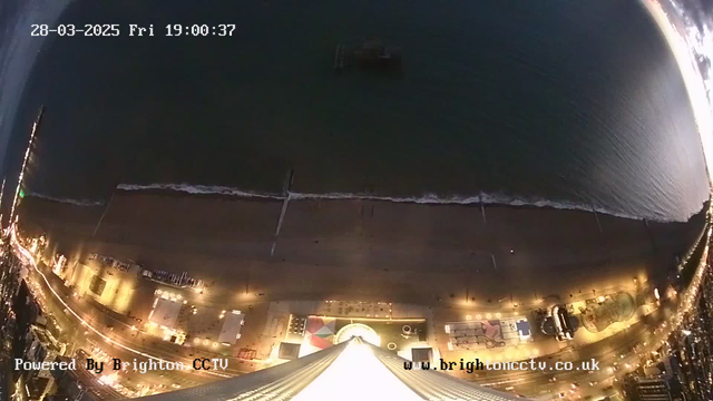 Aerial view of a beach at dusk, with the shoreline visible along the bottom of the image. The water is dark and slightly reflective, showing small waves. Brightly lit amusement rides and structures are lined up along the beach promenade, creating a colorful contrast against the sand. The lights from the attractions add warmth to the scene, with some shadows cast on the ground. The sky is transitioning from light to dark, indicating sunset. A digital timestamp is displayed in the top left corner.