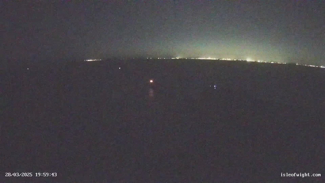 A dark scene depicting a distant horizon with faint lights. The sky transitions from dark to lighter shades near the horizon, suggesting twilight or dawn. Small points of light are scattered across the dark water, possibly indicating boats or buoys. The bottom of the image displays a timestamp in white text, along with a website URL.