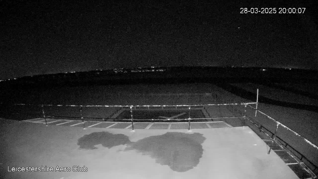 A dark scene captured by a webcam at night. In the foreground, there's a flat surface with a section that appears wet, likely reflecting some light. There is a railing at the edge of this surface, and the background features a large, open space with faint lights visible in the distance, indicating the presence of buildings or vehicles. The sky is predominantly dark, with a few stars visible. The timestamp at the top right shows the date and time as March 28, 2025, at 20:00:07. The bottom left corner displays the text "Leicestershire Aero Club."