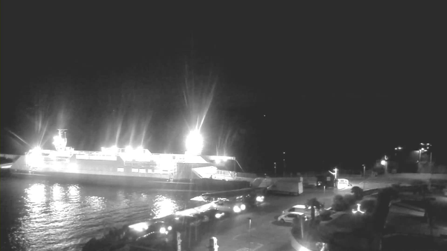 A nighttime scene showing a large ferry docked at a port, illuminated by bright lights. The water is calm, reflecting the lights. In the foreground, several vehicles, including trucks and cars, are parked near the dock. There are some structures visible on the right side, likely part of the port's infrastructure. The background appears dark, indicating it's nighttime.