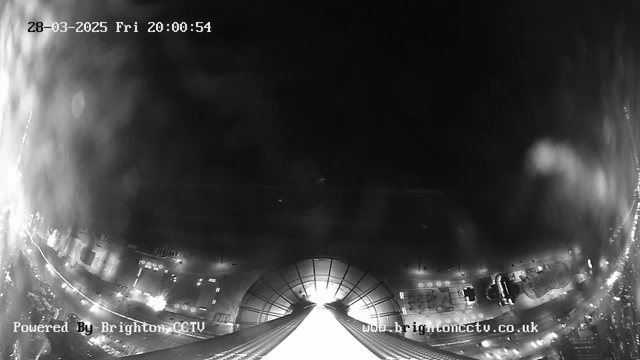 A black and white night scene captured from a webcam showing a view from a tall structure. At the bottom, there is a wide, illuminated pathway leading toward a bright light in the distance, likely indicating the ground or a structure below. Surrounding the pathway, there are faint outlines of buildings and streets, with some scattered lights appearing throughout the area, creating a sense of urban activity. The sky is dark and mostly empty, with a few clouds visible. The image is timestamped at 20:00:54 on March 28, 2025.