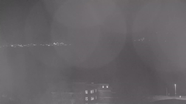 The image shows a dimly lit view captured by a webcam at night. In the foreground, a dark building is visible, featuring some lit windows. In the background, there are faint lights glimmering along a horizon, suggesting a distant cityscape or shoreline. The overall scene appears hazy and lacks clarity due to low light conditions.