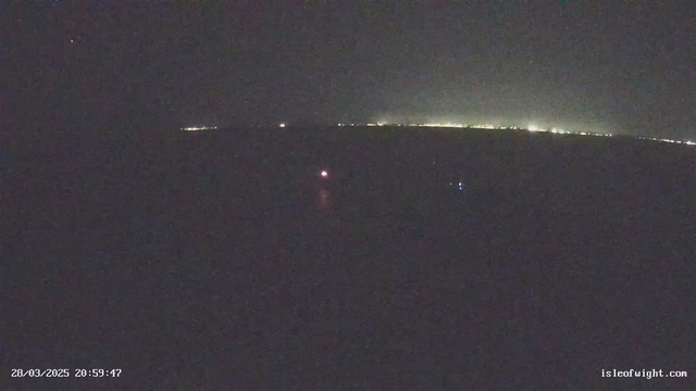 A dark scene captured by a webcam at night. The background shows distant city lights creating a faint glow along the horizon. In the foreground, there are two small, bright lights reflecting on dark water; one light is red and the other is blue. The image provides a sense of calm water with a mostly black sky. The time stamp in the lower corner indicates the date and time of the image.