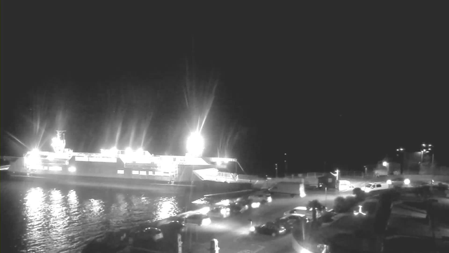 A brightly lit ferry is docked at a pier at night, reflecting light on the water. Several cars are parked and moving along a road adjacent to the pier. Streetlights illuminate the area, creating a contrast against the dark sky.
