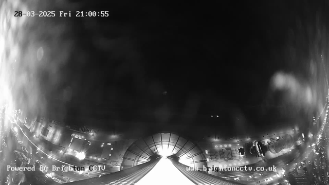 A black and white webcam image showing a view from a high location. The scene is mostly dark with some light appearing in the center, suggesting the presence of buildings and street lights below. The bottom of the image shows a sloped pathway leading to the light source, possibly indicating a structure or an entrance. The edges of the image have blurred elements, likely due to rain or condensation on the lens, obscuring some details. The date and time are displayed in the top left corner, while the words “Powered By Brighton CCTV” are in the lower left.