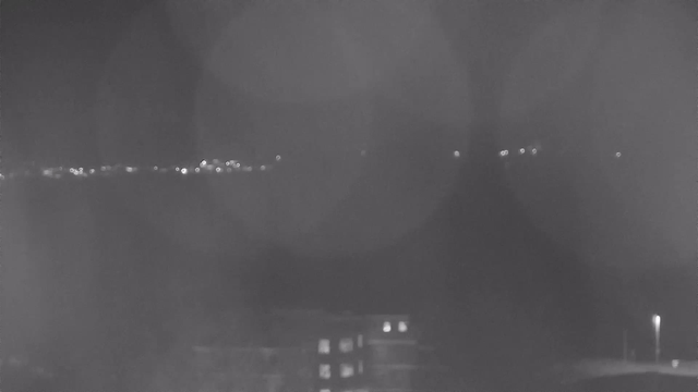 A black and white image showing a distant cityscape at night. Faint lights are visible, suggesting buildings and streetlights along the horizon. The foreground is dark, with some blurred spots indicating possible lens glare or moisture. Overall, the scene conveys a quiet, nighttime atmosphere.