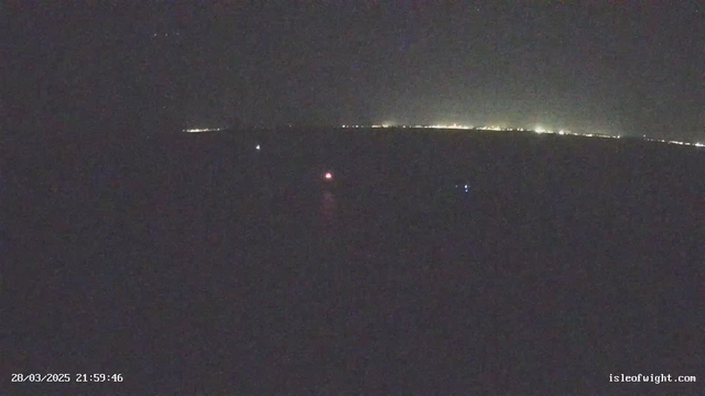 A dimly lit nighttime scene over water, with faint lights visible on the horizon. A few small, colored lights can be seen on the water. The overall atmosphere is dark, with variations of light scattered across the image, suggesting a coastal or maritime area.