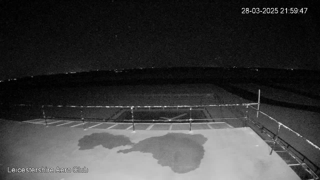 A dark scene captured from a webcam at the Leicestershire Aero Club. The image shows a flat surface with a water puddle reflecting some light, surrounded by a railing. The background is mostly dark, with faint lights visible in the distance, suggesting the presence of a horizon or landscape. The time stamp in the corner indicates the date and time of the capture. The overall atmosphere is nighttime with limited visibility.
