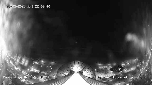 A black and white image taken from a high vantage point, showing a cityscape at night. The bottom of the image features a wide, curved structure that appears to be a building or roof. Below, streets are visible, illuminated by streetlights and car headlights creating a pattern of light. The sky is mostly dark, with some blurred spots possibly indicating light reflections or weather effects. The date and time, as well as a small watermark, are displayed in the corners.