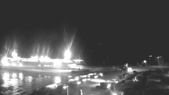 A nighttime scene featuring a large ship docked at a port, illuminated by bright lights. The water reflects the lights and is slightly rippling. Several vehicles are visible on a road near the dock, with headlights shining. People are seen on the edge of the dock, possibly observing the ship. The background is dark, emphasizing the lights from the ship and vehicles.