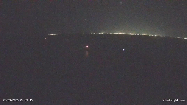 A dark scene captured at night, featuring a vast expanse of water under a starry sky. In the distance on the horizon, faint lights indicate land, suggesting the presence of a coastline. Closer to the foreground, a small bright light appears on the water, likely a boat or buoy. The overall atmosphere is quiet and serene, with a predominance of dark shades punctuated by the distant illuminations.