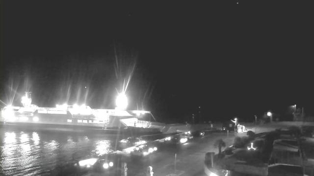 A nighttime scene at a harbor featuring a brightly lit ferry docked at the pier. The water reflects the lights from the ferry and nearby vehicles. Several cars are parked along the edge of the harbor, and the area is illuminated by streetlights. There are dark shapes of trees and structures in the foreground. The sky appears mostly dark with a few scattered lights.