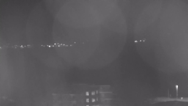 A dark, nighttime scene featuring a view of distant city lights along a horizon. The lights appear as small, bright dots against a dark background, with some faint reflections suggesting the presence of water below. In the foreground, an indistinct building is visible, with a few windows that exhibit light, but most of the image is shrouded in darkness. There are hazy circular shapes, possibly indications of camera lens reflections.