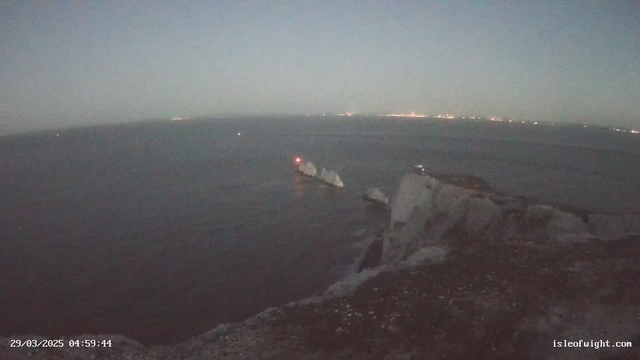 A coastal view at dawn, featuring a calm sea with a few low, rocky formations emerging from the water. A small lighthouse with a red light is visible on one of the rocks. In the distance, faint lights from a shoreline can be seen, suggesting a nearby town. The sky is lightening, transitioning from dark blue to a lighter hue. The atmosphere is tranquil, with gentle waves lapping at the rocks.