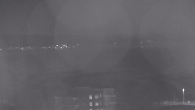 A dimly lit night scene captured by a webcam shows distant lights sparkling on the water's surface. The foreground features a building with a few illuminated windows, while the background includes a hazy view of water and faint outlines of further structures along the shoreline. The overall tone is dark and blurred, with shadows appearing throughout the image.