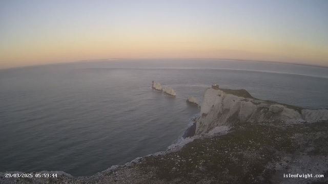 A rocky coastline with white cliffs is visible on the right side of the image. In the water, there are several large rocks, one of which has a red and white lighthouse. The sea is calm and reflects the pastel colors of the early morning sky, which transitions from peach to blue as it meets the horizon. A small building sits atop the cliff on the right. The date and time are displayed in the bottom left corner.