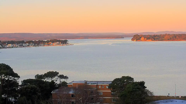 A calm waterway stretches out towards the horizon, reflecting soft hues of orange and pink from the sunset. On the left side, a small marina with several boats is visible, surrounded by buildings and trees. In the distance, an island with greenery and a structure can be seen, framing the natural landscape. The scene conveys tranquility, with hills visible beyond the water in the background.