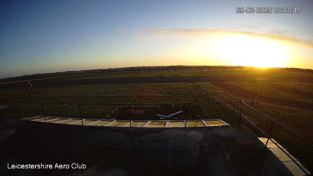 A scenic view of a clear sky at sunrise, with a gradient of warm colors from yellow to orange on the horizon. Below, there is a grassy airfield marked by white lines and a small airplane outline on the ground. The foreground features a railing, suggesting a vantage point, while the background extends into open fields and distant trees under the morning light. The image captures a tranquil and expansive landscape.