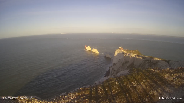 A coastal scene depicting white chalk cliffs towering over a calm sea. Several distinctive rock formations can be seen in the water, with a lighthouse on one of the rocks illuminated by morning light. The sky above is mostly clear with soft, pastel hues, indicating dawn. Shadows stretch across the foreground, highlighting the texture of the cliffside. A small boat is visible in the distance on the water.