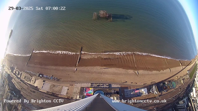 The image shows a wide view of a coastline with a sandy beach meeting the ocean. Gentle waves lap against the shore, with a wooden pier partially visible in the water. To the left, remnants of an old pier can be seen jutting out, and along the beach, there are various structures, possibly shops or amusement rides, with colorful markings. The image is taken from a high vantage point, looking down towards the beach. The date and time are displayed in the upper left corner.