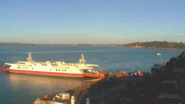 A white and red ferry is docked at the waterfront, with its ramp lowered. The calm water reflects the blue sky, while hills can be seen in the distance. Nearby, small boats float, and a few structures line the shore, including some vehicles parked and a section of a pathway. The overall scene is peaceful, with no clouds in the sky.