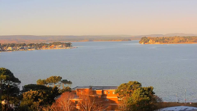 A scenic view of a calm body of water framed by trees and buildings. In the foreground, there are patches of green foliage and an orange-brick building. The water reflects soft sunlight, and in the distance, there is a small harbor with boats and a tree-lined shoreline. Beyond the water, rolling hills are visible under a clear sky. The overall atmosphere conveys a peaceful, natural setting.