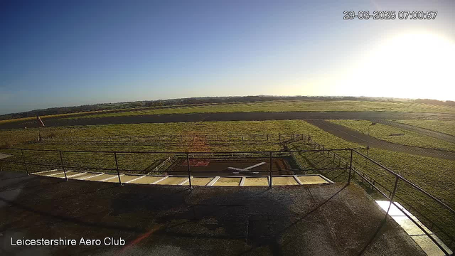 A clear blue sky is visible with no clouds. In the foreground, there is a paved area with a small helicopter landing pad marked by a white "X." Surrounding the landing pad are green grassy fields that extend towards the horizon. A faint outline of trees is seen in the distance. A flagpole stands to the left of the image, and a runway is visible on the right, leading across the fields. The time stamp shows the image was taken at 07:00:57 on March 29, 2025.