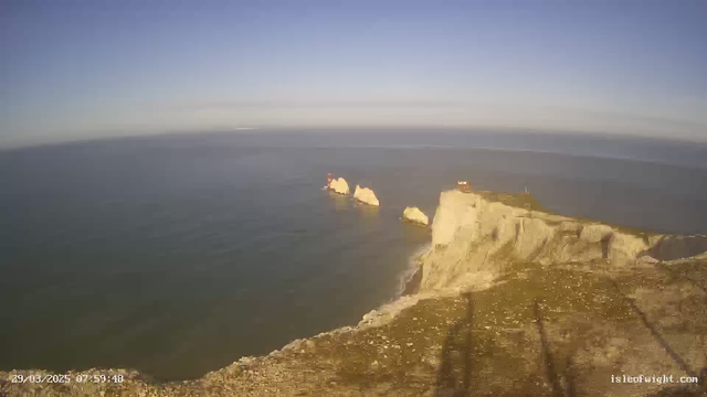 A panoramic view of a coastline featuring steep, rugged cliffs meeting the ocean. The water is calm and reflects the clear blue sky, which has a few wispy clouds. In the distance, some rock formations extend into the sea, with a small red structure visible on one of the rocks. The foreground shows the grassy top of the cliff, with some shadows cast by an unseen object.
