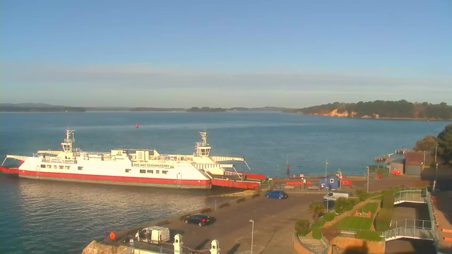 A white and red ferry is docked at a terminal next to calm blue waters. The boat has several visible windows and a noticeable bridge. In the foreground, there are some cars parked on a paved area, with a few palm trees and landscaped greenery nearby. On the opposite side, a wooden jetty extends into the water, and the background features a hilly shoreline with trees. The sky is clear and bright, indicating a sunny day.
