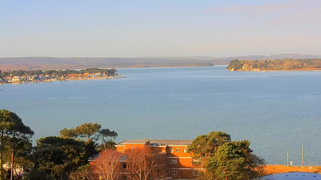 A tranquil coastal scene shows a wide, calm waterway surrounded by trees and buildings. In the foreground, there is vegetation and a low building with a reddish exterior. The background reveals a harbor with boats docked and a distant shore lined with trees, hills, and a gentle blue sky. The light is soft, indicating a clear, sunny day.