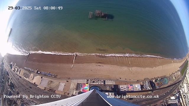 Aerial view of a beach and coastline. The sandy beach stretches along the bottom of the image with gentle waves lapping at the shore. A wooden pier juts out into the water from the beach area, while several people walk along the beach. In the background, the sea appears calm and blue. Beneath the beach, there are colorful structures, possibly amusement park rides, visible along the promenade. The image includes a timestamp indicating the date and time, with a logo for Brighton CCTV at the bottom.