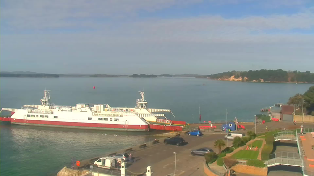 A large white ferry with red accents is docked at a harbor, surrounded by calm water. In the background, green hills are visible under a light blue sky with some clouds. A few boats can be seen in the distance, and along the shore, there is a parking area with parked vehicles and palm trees. Additionally, a red buoy floats in the water nearby.