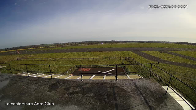 A view from a webcam looking out over a grassy field at an aero club. In the foreground, there is a small helipad marked with a white cross surrounded by a gravel area. A railing borders the edge of a building at the bottom of the image. The background features a wide, open space with a tarmac runway visible, leading into a horizon with patches of blue sky and scattered clouds. The overall scene is bright and sunny, indicating clear weather.