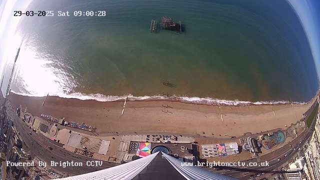 Aerial view of a beach with a wide expanse of sand and ocean. The water is calm with gentle waves lapping at the shore. Near the shoreline, a few people are walking. A colorful beach area with umbrellas and beach equipment is visible on the sand. In the distance, a partially submerged structure can be seen in the water. The beach is bordered by a lively promenade with various buildings and attractions. The sky is clear with some reflections on the water. A timestamp indicating the date and time is displayed in the top left corner.