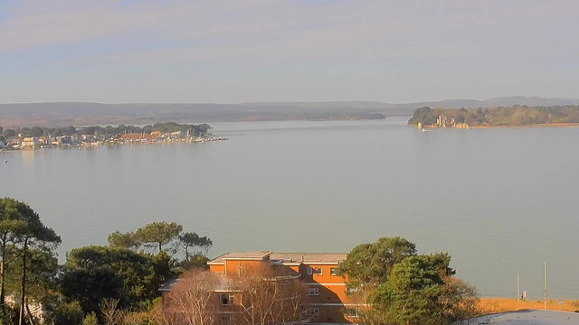 A serene landscape featuring a calm body of water reflecting a clear blue sky with soft clouds. On the left side, there is a small harbor with boats and a cluster of buildings, while the right side showcases a distant island with a castle-like structure. In the foreground, there are green trees and an orange-brick building. The overall scene conveys a peaceful and picturesque view of the waterfront.