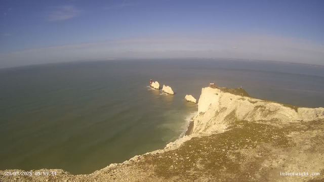 A coastal view featuring white chalk cliffs at the right side, leading into a calm blue sea that stretches to the horizon. In the distance, a lighthouse stands on a rocky outcrop, while two smaller rocks are visible in the water. The sky is clear with a few wispy clouds. The bottom of the image shows rocky terrain with sparse grass. The scene conveys a serene, natural landscape.
