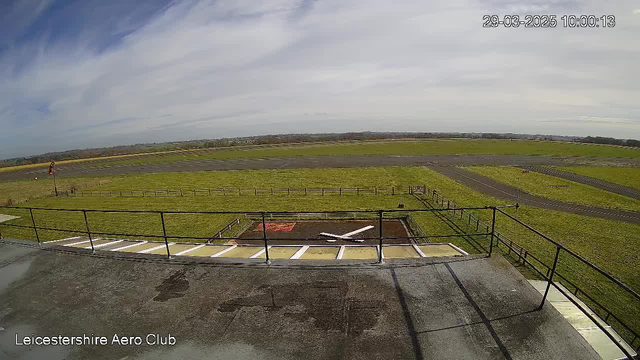 A wide view of a grassy airfield taken from a high vantage point, featuring a small section of a flat rooftop in the foreground. There is a white cross painted on the ground, possibly for aircraft landing guidance. The landscape is mostly flat with a few scattered bushes and a distant tree line under a partly cloudy sky. A wooden fence runs along the edge of the airfield, and a runway is visible in the distance. The bottom left corner contains the text "Leicestershire Aero Club."