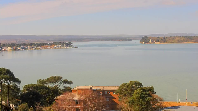 A scenic view of a calm body of water, with a marina lined by colorful boats and buildings on the left side. In the background, lush green hills fade into the distance under a clear blue sky, with soft clouds above. The shoreline features some trees and a few large structures nestled among them. The overall scene conveys tranquility and natural beauty.