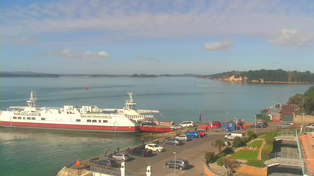 A view of a calm body of water with a large ferry docked at a pier. The ferry is white with a red stripe and has vehicles boarding. In the foreground, a parking area is filled with several cars. There are people gathered near the water's edge. On the right side, a red building and a dock area are visible, along with a landscaped area featuring grass and pathways. The sky is mostly clear with a few clouds, and the shoreline in the background is lined with green trees and rocky outcrops.
