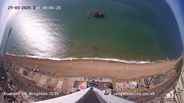 Aerial view of a beach and coastline on a sunny day. The beach has a sandy shoreline with gentle waves lapping at it. A pier with a structure partially submerged in the water can be seen. There are several people strolling along the beach and various colorful beach huts and structures visible. The image captures the vibrant blue of the sea and the reflection of sunlight on the water's surface, with the sky above being mostly clear. The bottom part of the image includes a glimpse of amusement rides and colorful patterns on the boardwalk leading to the beach area.