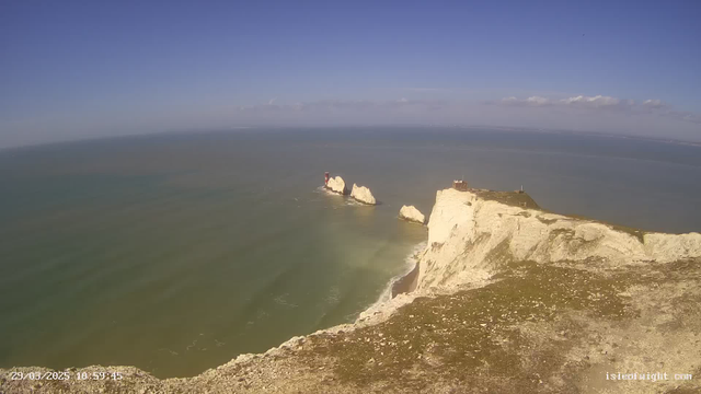 A panoramic view of a coastline featuring steep, white chalk cliffs that drop into the sea. In the distance, there are three prominent rock formations partially emerging from the water. A red and white lighthouse is positioned on one of the rocks, while a small structure sits on top of the cliff to the right. The sky above is clear with some clouds, and the sea exhibits a mix of blue and green hues. The foreground is rocky with sparse vegetation.