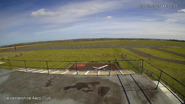 A view from a webcam at the Leicestershire Aero Club, showing a large, open green field with a clear blue sky above. In the foreground, there's a flat rooftop with railings and some water marks. The runway is visible, leading into the distance, with a small red marker and a white cross on a patch of brown ground, surrounded by fencing. The sky is partly cloudy, suggesting a sunny day.