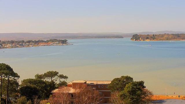 A scenic view of a calm body of water, with gentle waves reflecting shades of blue and green. In the foreground, there are trees with varying shades of green, and a building with a reddish exterior is partially visible. The midground features small boats scattered across the water, while in the distance, a shoreline with houses and a marina can be seen. The background comprises rolling hills under a clear sky.