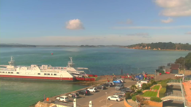 A scenic view of a harbor on a clear day. A large white ferry is docked at the pier, with its name partially visible. The water is a vibrant turquoise and reflects the blue sky, dotted with a few clouds. In the foreground, there are several parked cars and people gathered near the ferry entrance, while lush green grass and landscaped areas are visible on the right. The coastline in the background shows a tree-lined shore.