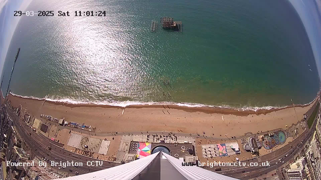 Aerial view of a beach with a broad expanse of golden sand meeting teal waves. The shoreline features a pier extending into the water and several scattered beachgoers. In the foreground, a park area displays colorful designs on the ground, with several amusement rides visible. The sky is mostly clear with some patches of blue, and the sunlight glistens on the surface of the water. In the top left corner, the date and time are displayed.