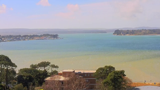 A scenic view of a calm body of water, with gentle waves reflecting shades of blue and green. In the background, there are several boats and a small harbor lined with colorful structures. Across the water, faint hills rise, partially obscured by a light haze. In the foreground, trees and a building with a flat roof can be seen, adding depth to the landscape. The sky is mostly clear with a few soft clouds scattered throughout.