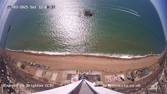 Aerial view of a sandy beach along a coastline, with gentle waves lapping at the shore. In the foreground, a tall structure extends downward, with the point of view looking straight down. The beach is dotted with tiny figures of people, and colorful beach umbrellas can be seen. To the right, an amusement area features a swimming pool and various attractions. In the distance, a pier extends into the water. The sky above is clear with a few wispy clouds. The image includes a timestamp in the upper left corner.