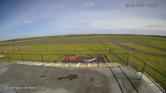 A wide view of an airfield with a grassy landscape under a clear blue sky. There are a few white clouds visible. In the foreground, a metal railing encloses what appears to be a flat roof or terrace. Beneath the railing, there is a painted runway area marked with an 'X' and white lines. In the distance, the airfield stretches out with a runway and several grassy patches, bordered by low fences. A small red flag stands on the runway, indicating wind direction. The date and time are displayed in the top right corner, showing March 29, 2025, at noon.